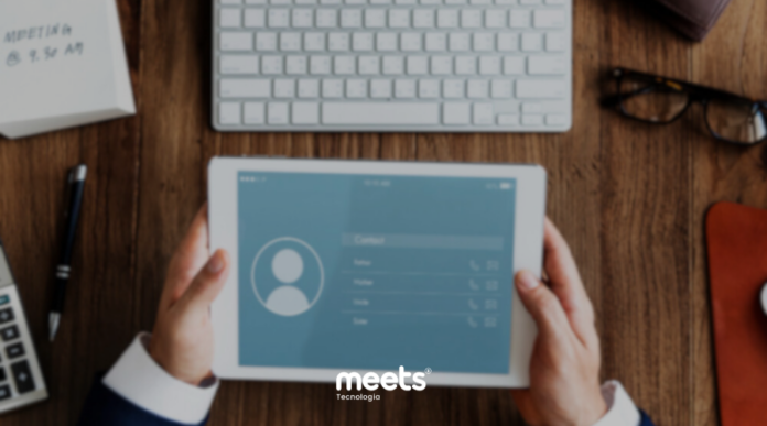 Visão aérea de uma mesa de trabalho de madeira com um teclado, óculos, caneta e calculadora. No centro, mãos seguram um tablet exibindo uma interface de contatos desfocada. Abaixo, o logotipo 'meets Tecnologia'