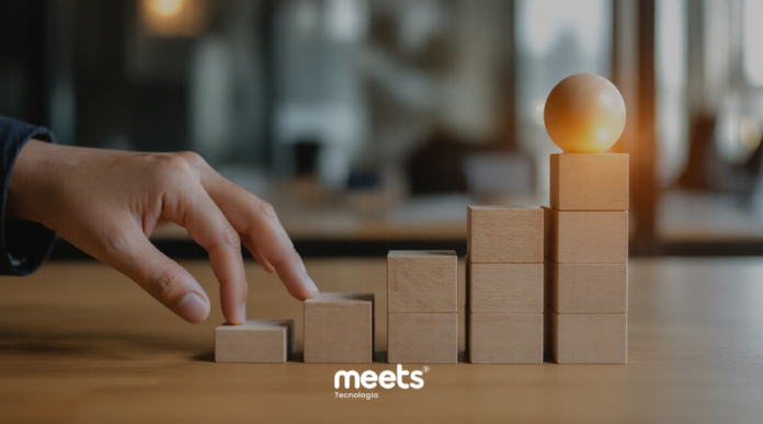Mão posicionando blocos de madeira em formato de escada sobre uma mesa, com uma esfera no topo, representando crescimento e evolução estratégica em um ambiente corporativo desfocado.