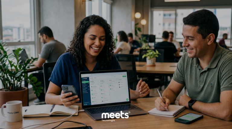 Dois profissionais sorridentes trabalhando em uma mesa de escritório moderno. Uma mulher segura um smartphone e aponta para um laptop que exibe uma tela de sistema de CRM com funil de vendas, enquanto um homem faz anotações em um caderno.